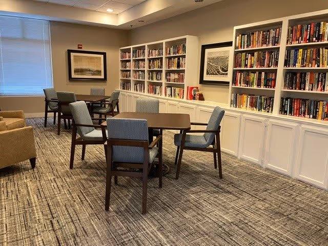 A cozy reading and seating area in a senior living facility featuring several wooden tables with cushioned chairs, bookshelves filled with books along the wall, framed artwork, and large windows with blinds letting in natural light.