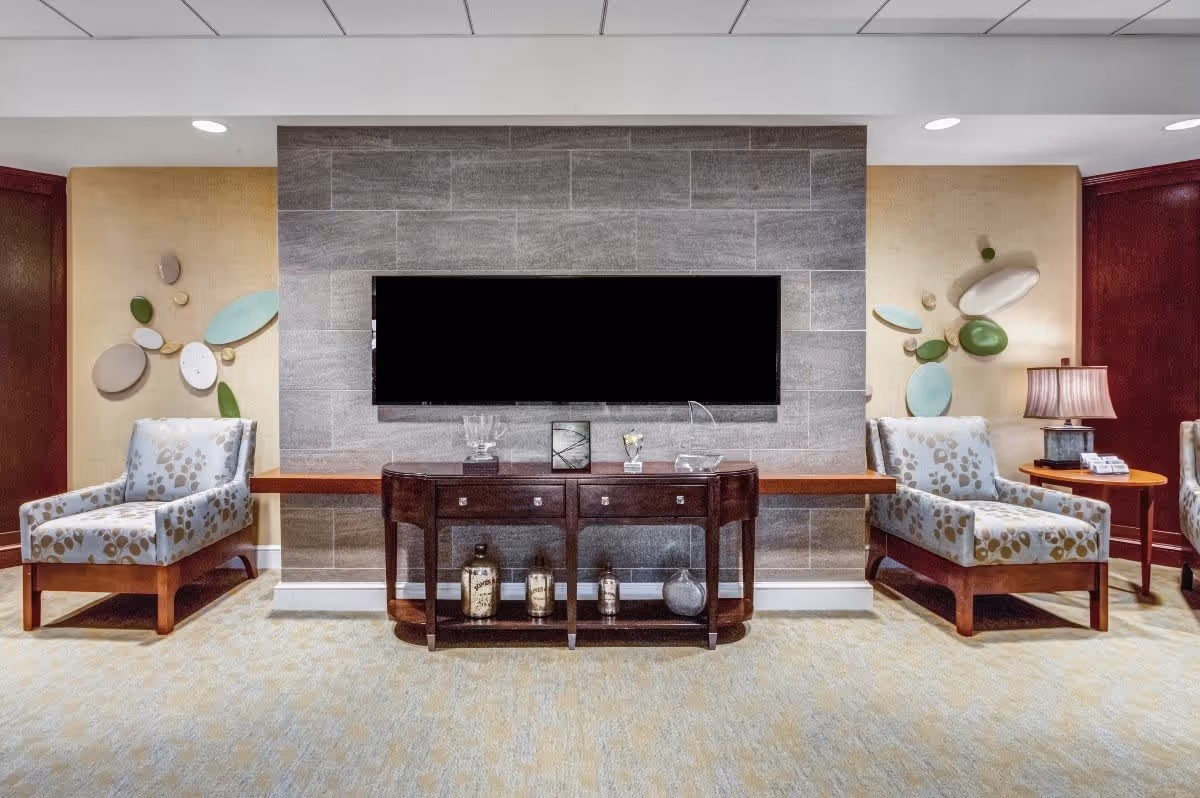 A cozy living room area with two patterned armchairs on either side of a wooden console table. The console table is placed against a gray tiled wall with a large flat-screen TV mounted above it. Decorative items are displayed on the console table and on the wall behind the chairs. A small round side table with a lamp is next to the right armchair.