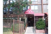 Entrance to Grand Manor Nursing & Rehabilitation Center with a maroon awning displaying the facility's name above a set of stairs, surrounded by a metal fence and greenery.