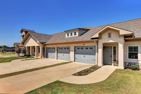Exterior view of a senior living facility building with multiple garage doors and a front entrance door, surrounded by a concrete driveway and green lawn under a clear blue sky.