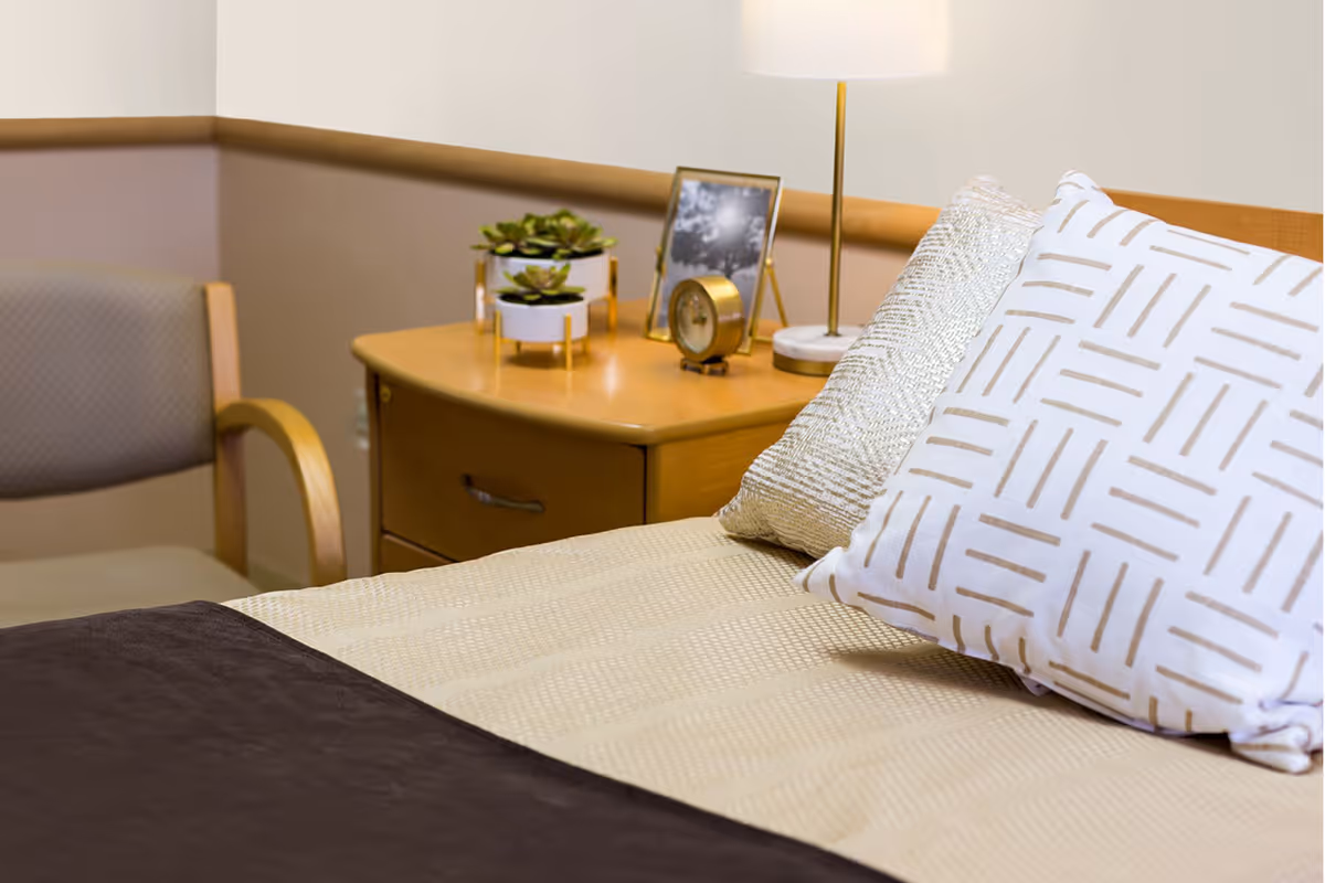 A neatly made bed with two decorative pillows, a beige bedspread, and a dark brown blanket. Next to the bed is a wooden nightstand with a small lamp, a framed photo, a clock, and two small potted succulent plants. A cushioned chair with wooden armrests is positioned near the nightstand against a beige and light brown wall.