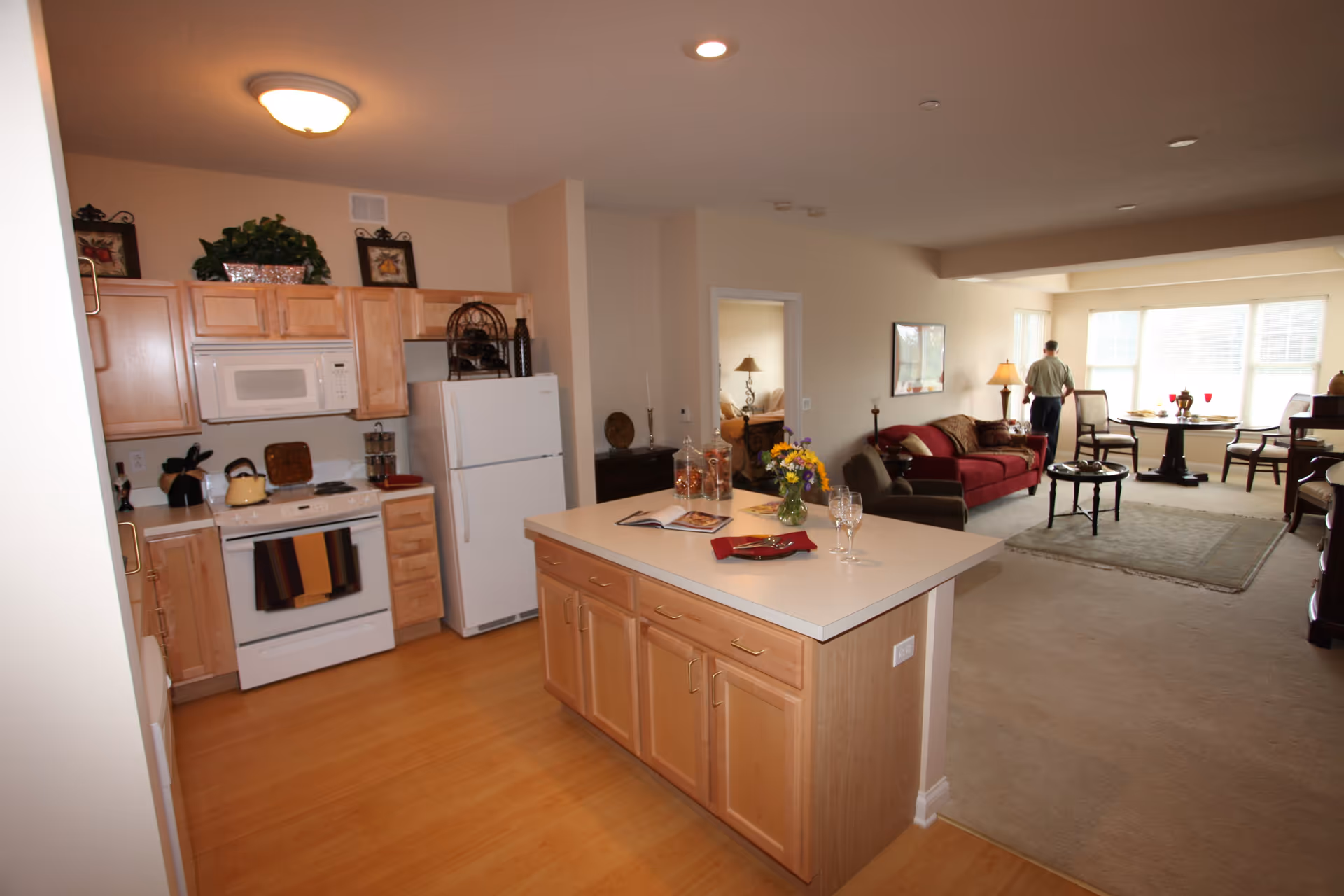Interior view of a senior living facility apartment showing a kitchen with wooden cabinets, white appliances including a microwave, stove, and refrigerator, and an island countertop with a vase of flowers and two wine glasses. Beyond the kitchen is a living room area with a red sofa, a coffee table, and a dining table near large windows. A person is standing near the windows looking outside.