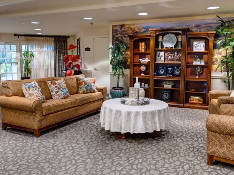 A cozy living room area with a brown patterned sofa and armchair, both adorned with floral cushions. A round table covered with a white tablecloth sits in the center, decorated with vases and a small sign. Behind the table is a wooden bookshelf filled with decorative items, books, and a stereo system. The room has patterned carpet flooring, potted plants, and a large window with sheer curtains allowing natural light to enter.