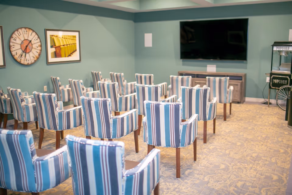 A small theater room with multiple striped upholstered chairs arranged in rows facing a large flat-screen TV mounted on a green wall. There is a popcorn machine on the right side and a large clock and framed picture on the left wall.