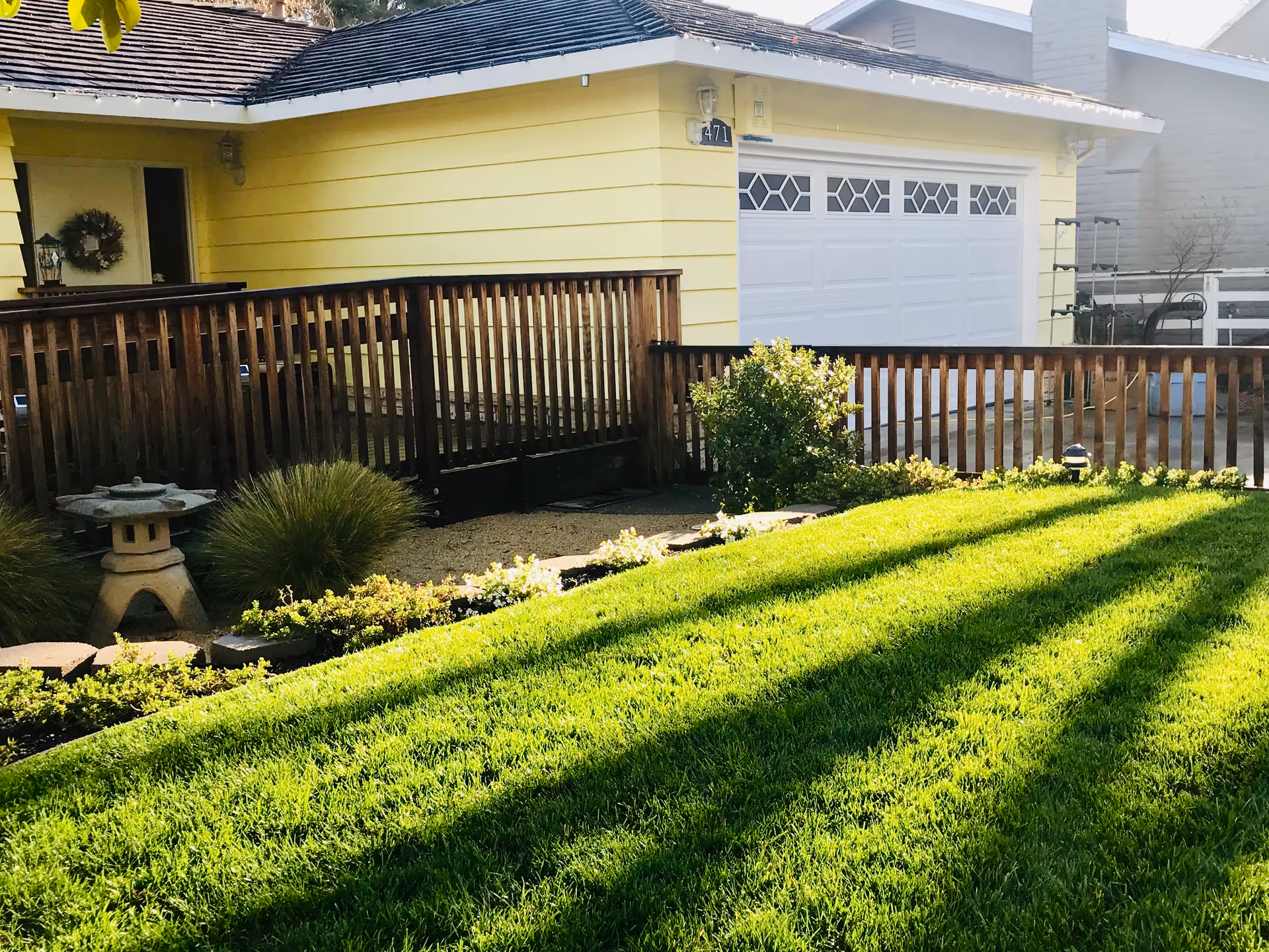 A well-maintained front yard with lush green grass, a wooden fence, and a yellow house with a white garage door in the background. There are some bushes and a small stone lantern near the fence.