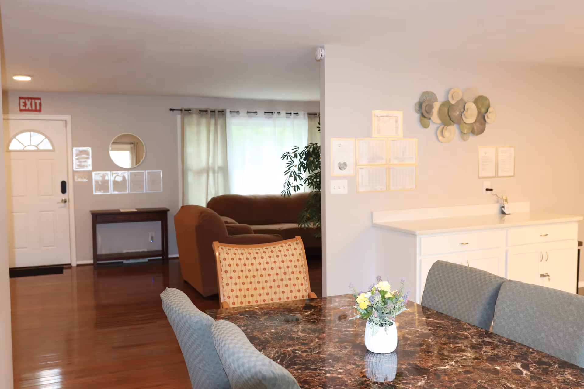 Dining area with a marble table and vase in the foreground, upholstered chairs, a countertop and cabinets to the right, and a living room with couches in the background.