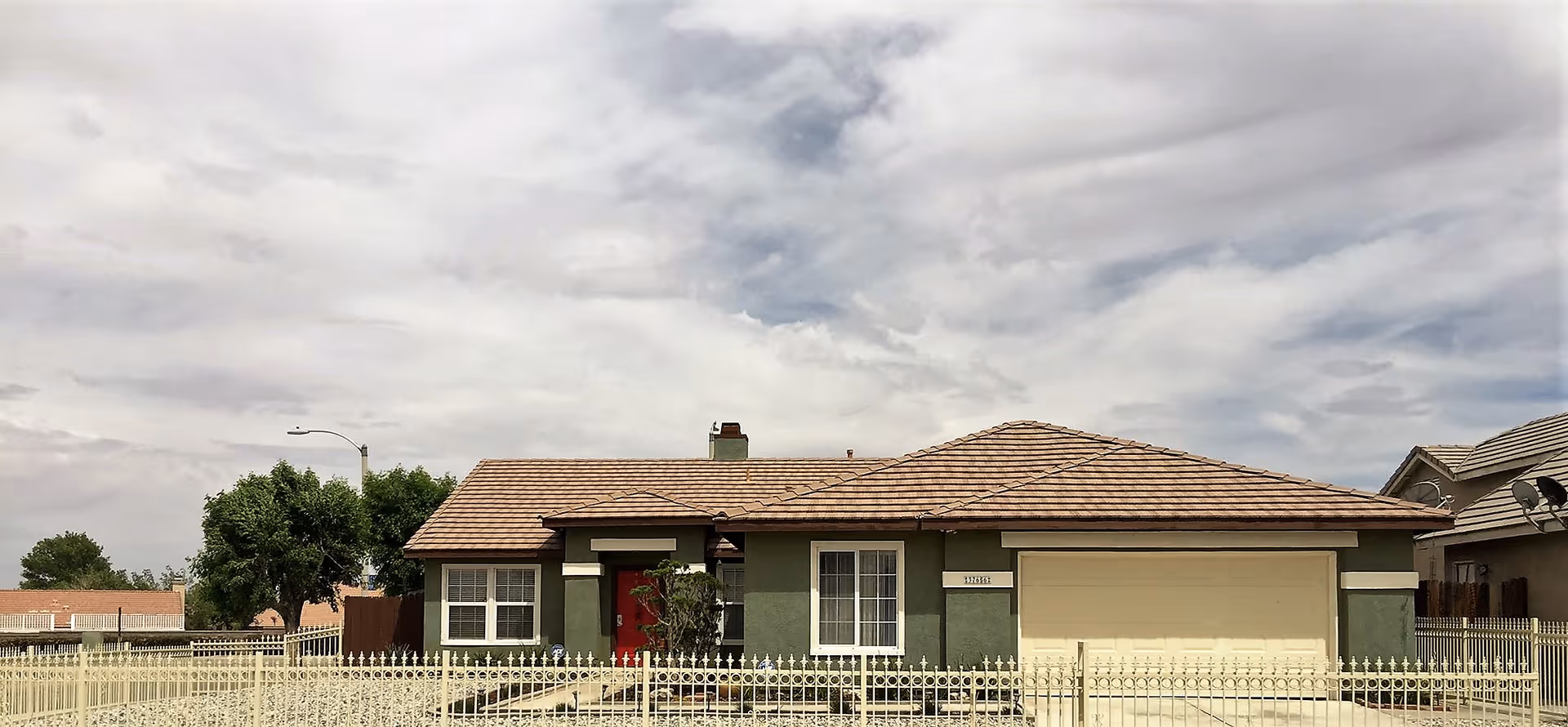 Single-story residential building with a tiled roof, green exterior walls, a red front door, and a two-car garage. The house is surrounded by a white metal fence and there are trees and other houses visible in the background under a cloudy sky.