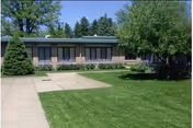One-story building with a green lawn, concrete walkways, and trees under a blue sky.