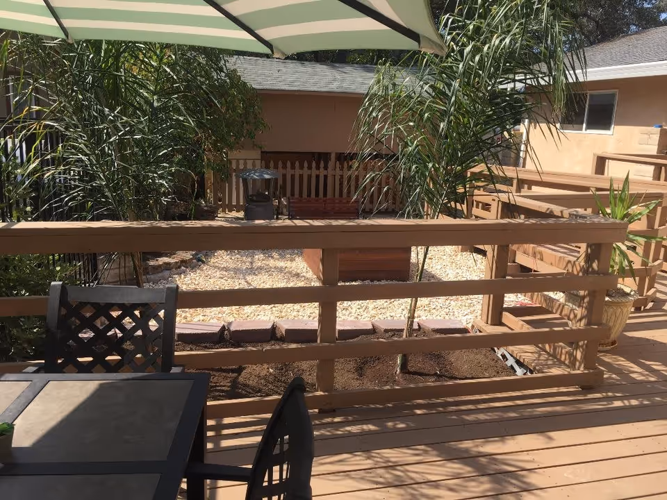 Outdoor patio area with a wooden railing, a table with chairs, potted plants, and an umbrella providing shade. In the background, there are palm trees, a small fenced garden area with gravel, and a building with beige walls and windows.