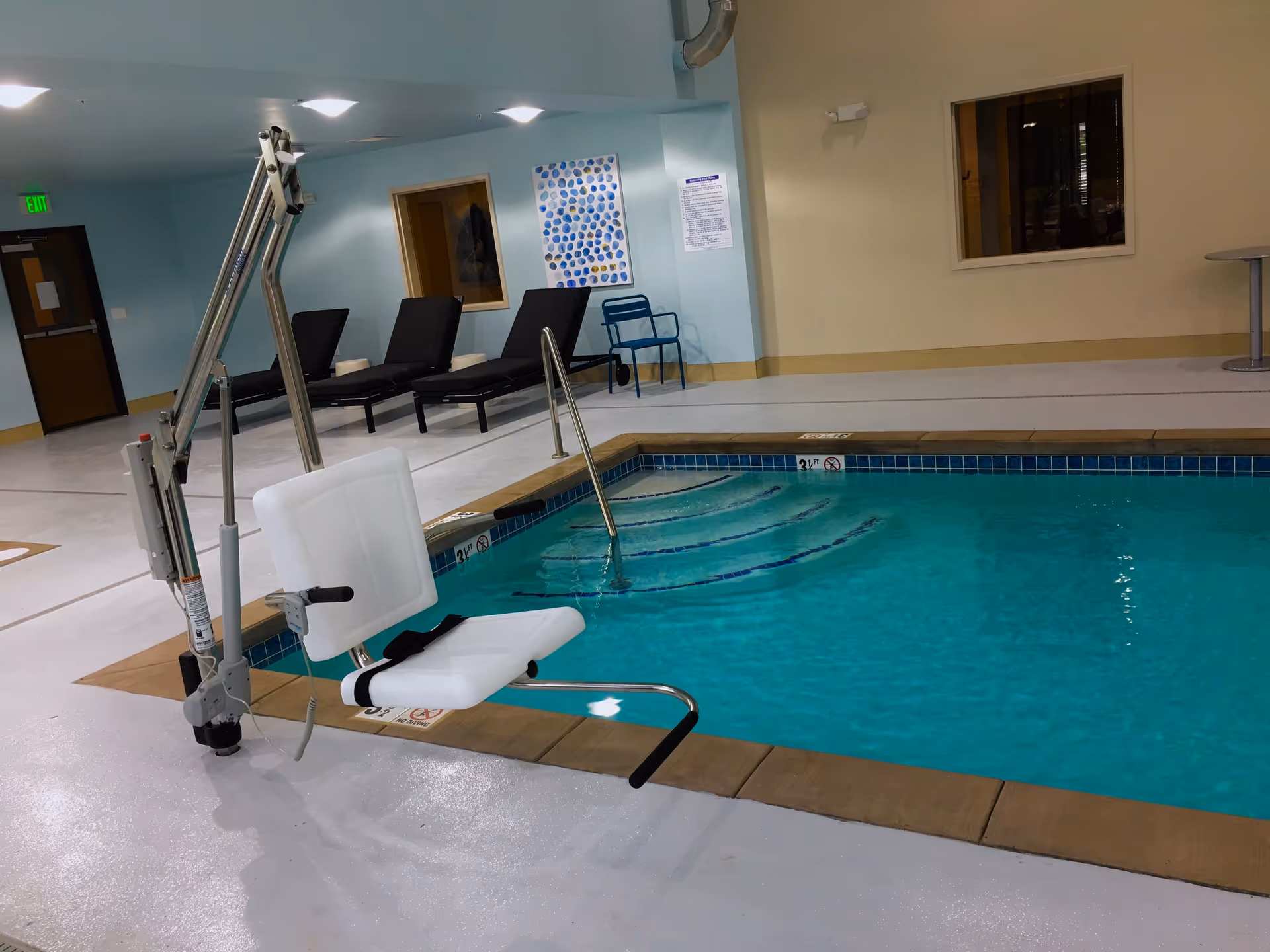Indoor swimming pool with a pool lift chair for accessibility. There are several black lounge chairs and a blue chair against the light blue and beige walls. The pool has steps leading into the water and depth markers indicating 3 feet.