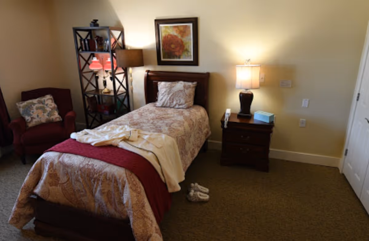 A cozy bedroom with a single bed covered in a patterned bedspread and a red throw blanket. A white robe and slippers are placed on the bed and floor. To the left of the bed is a red armchair with a decorative pillow, and behind it is a black metal shelving unit with lamps and decor. On the right side of the bed is a wooden nightstand with a lamp, a tissue box, and a phone. A framed floral picture hangs on the wall above the bed.