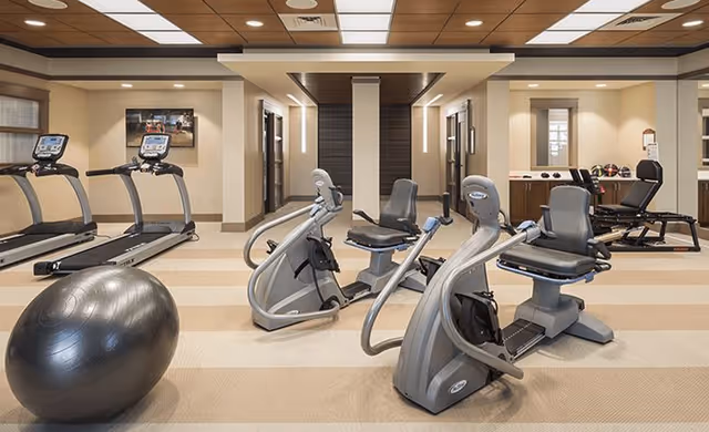 A well-lit fitness room featuring two treadmills, three recumbent exercise bikes, a large exercise ball, and various weights and exercise equipment along the walls. The room has a beige and brown color scheme with a tiled ceiling and recessed lighting.