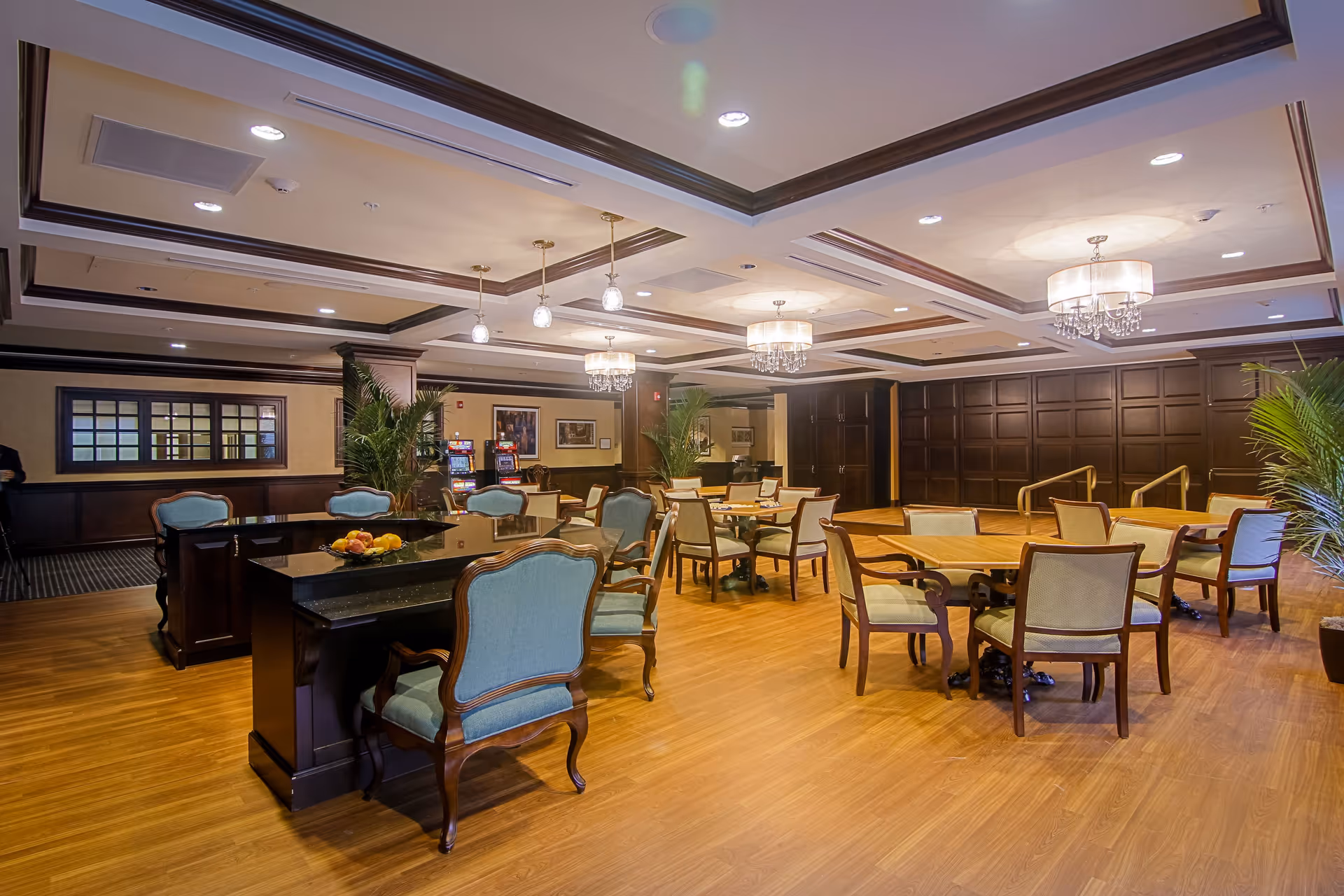 A spacious, well-lit common area with wooden flooring and elegant ceiling lights. The room features multiple tables and chairs with light blue upholstery, some arranged around a black granite-topped counter. There are large potted plants and framed artwork on the walls, creating a warm and inviting atmosphere.