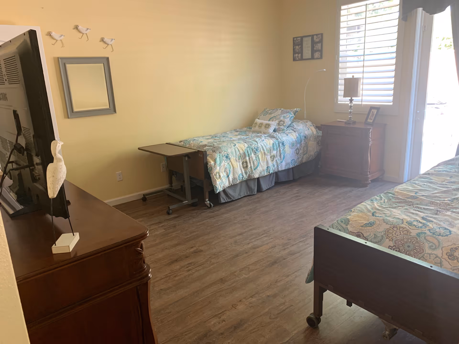 Sunny shared bedroom with two twin beds, wooden furniture, a TV on a dresser, and a bedside table by a window.