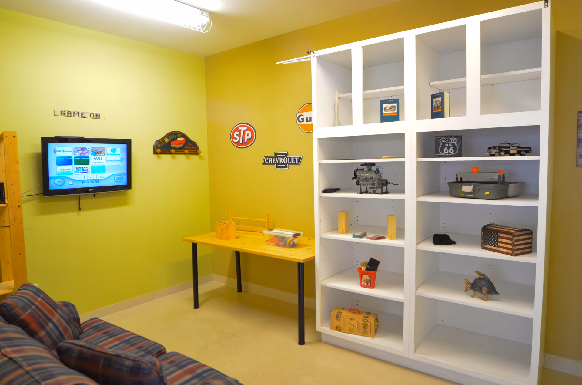 Activity room with a wall-mounted TV, a couch, a small yellow table and white shelving holding games and decor.