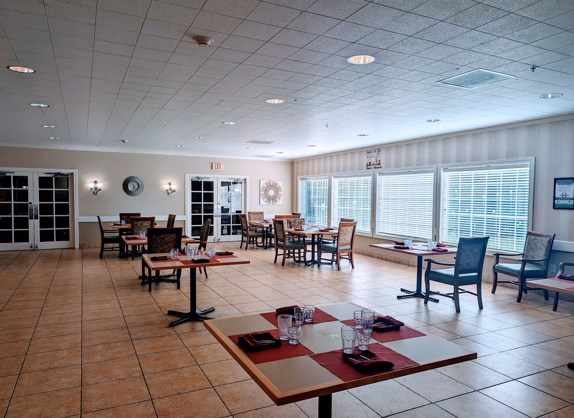 A bright dining room with several square tables set with glasses and napkins. The room has large windows with blinds, tiled floors, and beige walls. There are multiple chairs around the tables and wall decorations including a clock and framed art. Double doors are visible at the far end of the room.