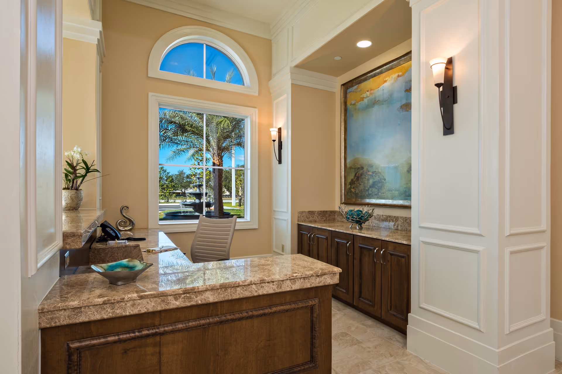 Reception area with a marble countertop desk, a white office chair, a window showing palm trees and a fountain outside, beige walls, wooden cabinets, wall sconces, and a large framed abstract painting.
