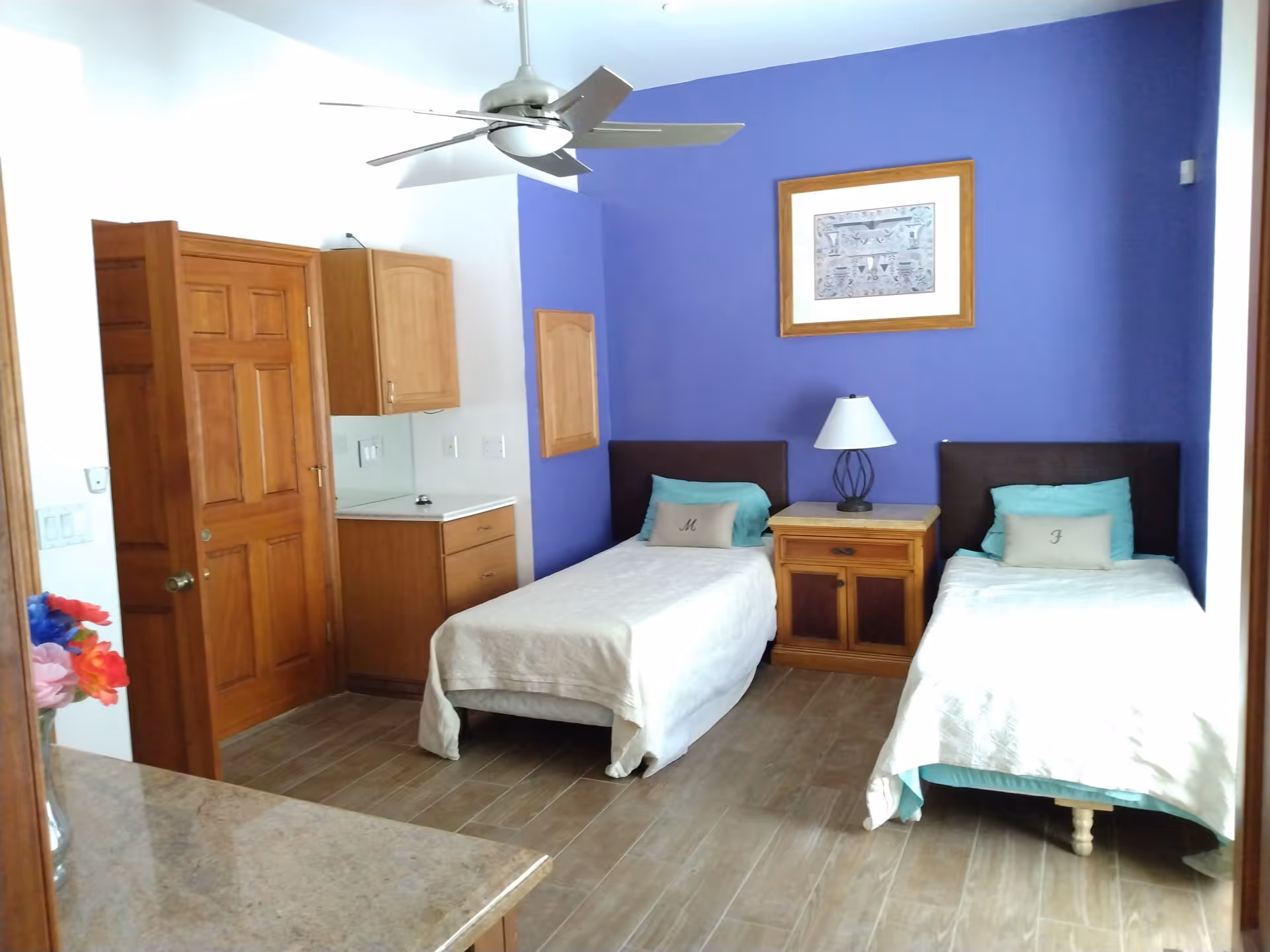 A bedroom with two single beds separated by a wooden nightstand with a lamp. The wall behind the beds is painted purple and has a framed picture. There is a ceiling fan above, wooden doors and cabinets on the left side, and a granite countertop in the foreground.