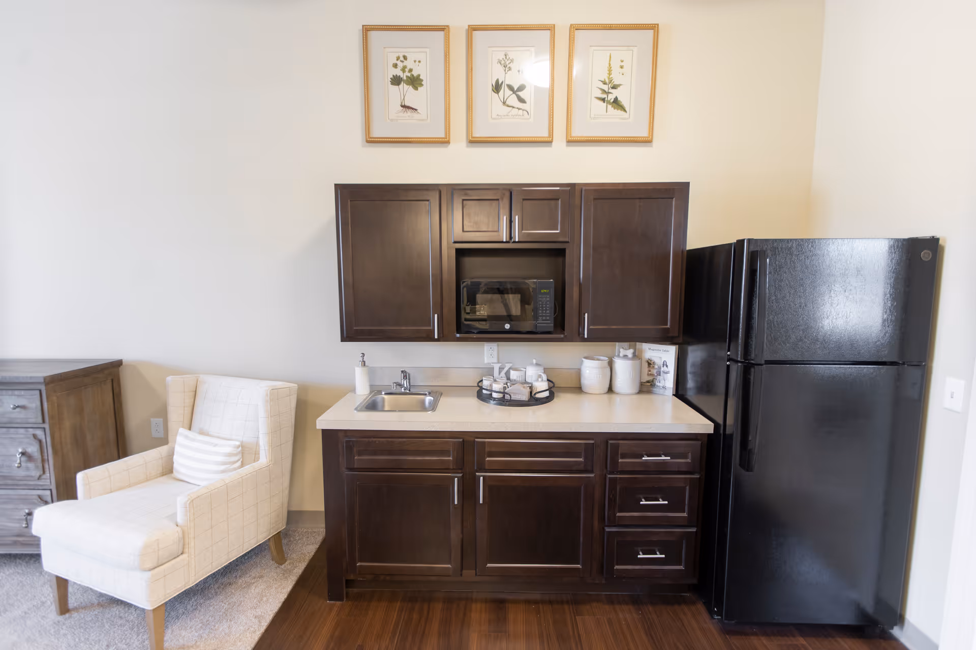 A small kitchenette area with dark wood cabinets, a black microwave, a black refrigerator, a small sink, and countertop with decorative jars and a tray. To the left of the kitchenette is a beige upholstered armchair with a striped pillow, and a wooden dresser. Three framed botanical prints hang on the wall above the cabinets.