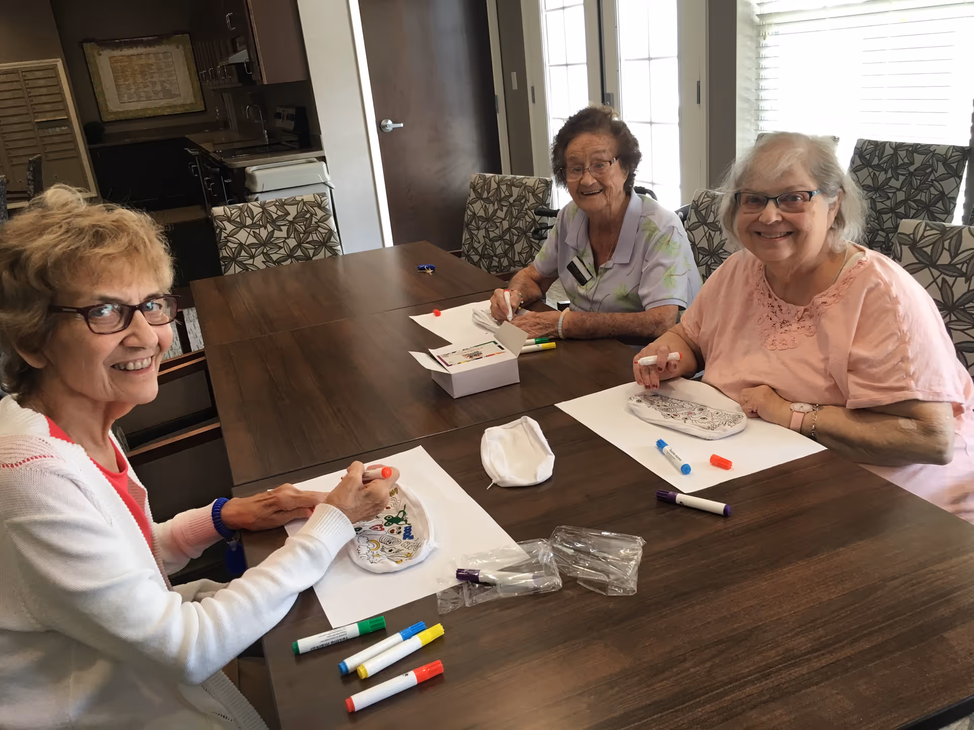 Three elderly women sitting around a wooden table in a well-lit room, smiling and engaging in a coloring activity with markers and coloring sheets in front of them.