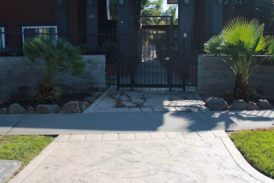 Entrance to a residential building with a black metal gate, flanked by low stone walls and small palm plants on either side. The pathway leading to the gate is paved and bordered by grass.