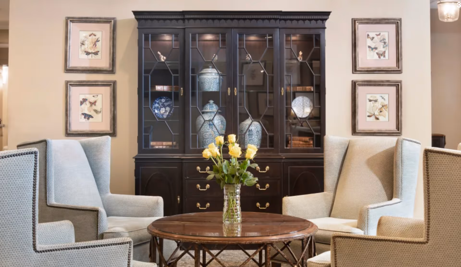 Cozy sitting area with upholstered armchairs around a round wooden table topped with a vase of yellow roses, set before a dark display cabinet and framed artwork.