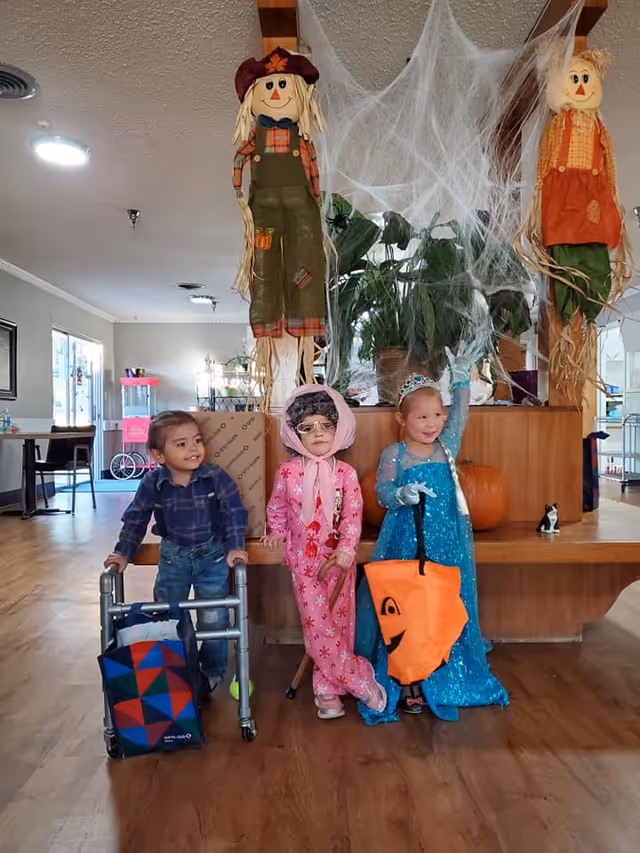 Three children dressed in Halloween costumes stand inside a room decorated with Halloween-themed items. One child is using a walker and holding a colorful bag, the second child is wearing a pink outfit with a bonnet and glasses, and the third child is dressed as a princess in a blue sparkly dress holding an orange pumpkin-shaped bag. Behind them are two scarecrow decorations hanging with fake spider webs and a large potted plant on a wooden counter. The room has wooden flooring and visible furniture in the background.