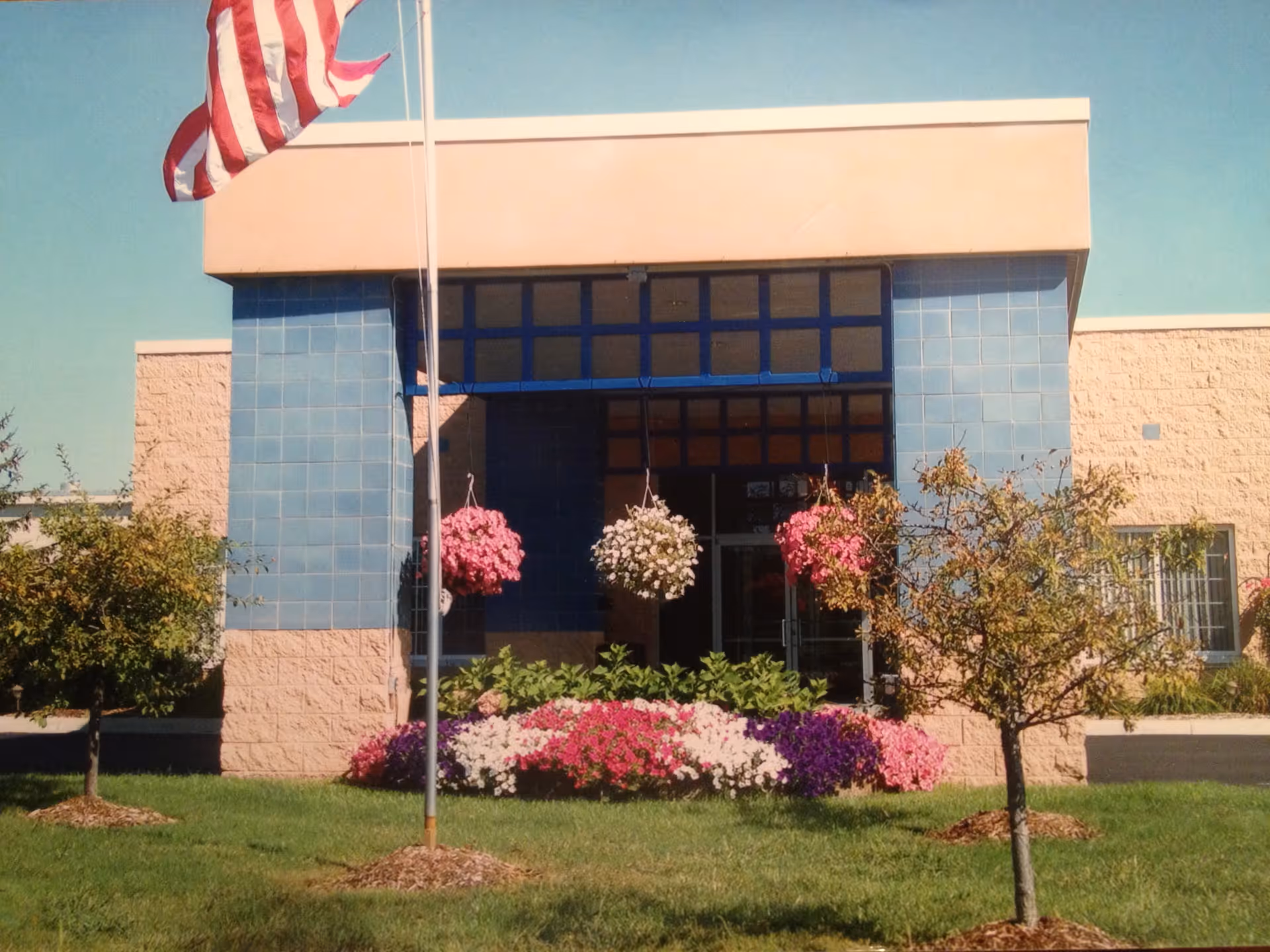 Front entrance with an American flag, blue-tiled entryway, hanging flower baskets, and landscaped flower beds.