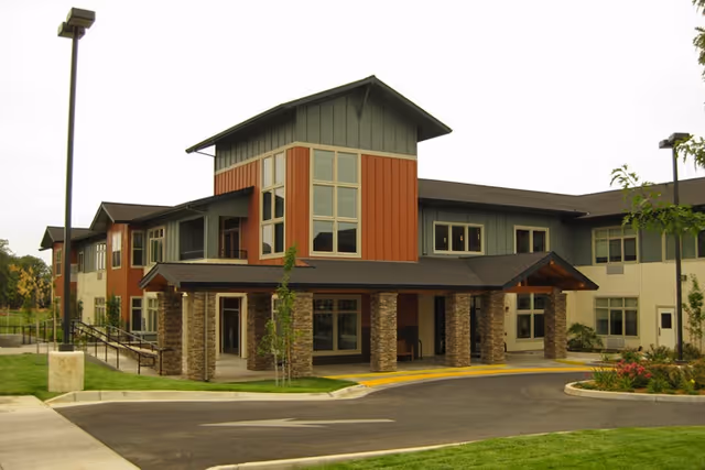 Modern two-story assisted living building with a covered entrance supported by stone columns, driveway, and landscaped grounds.
