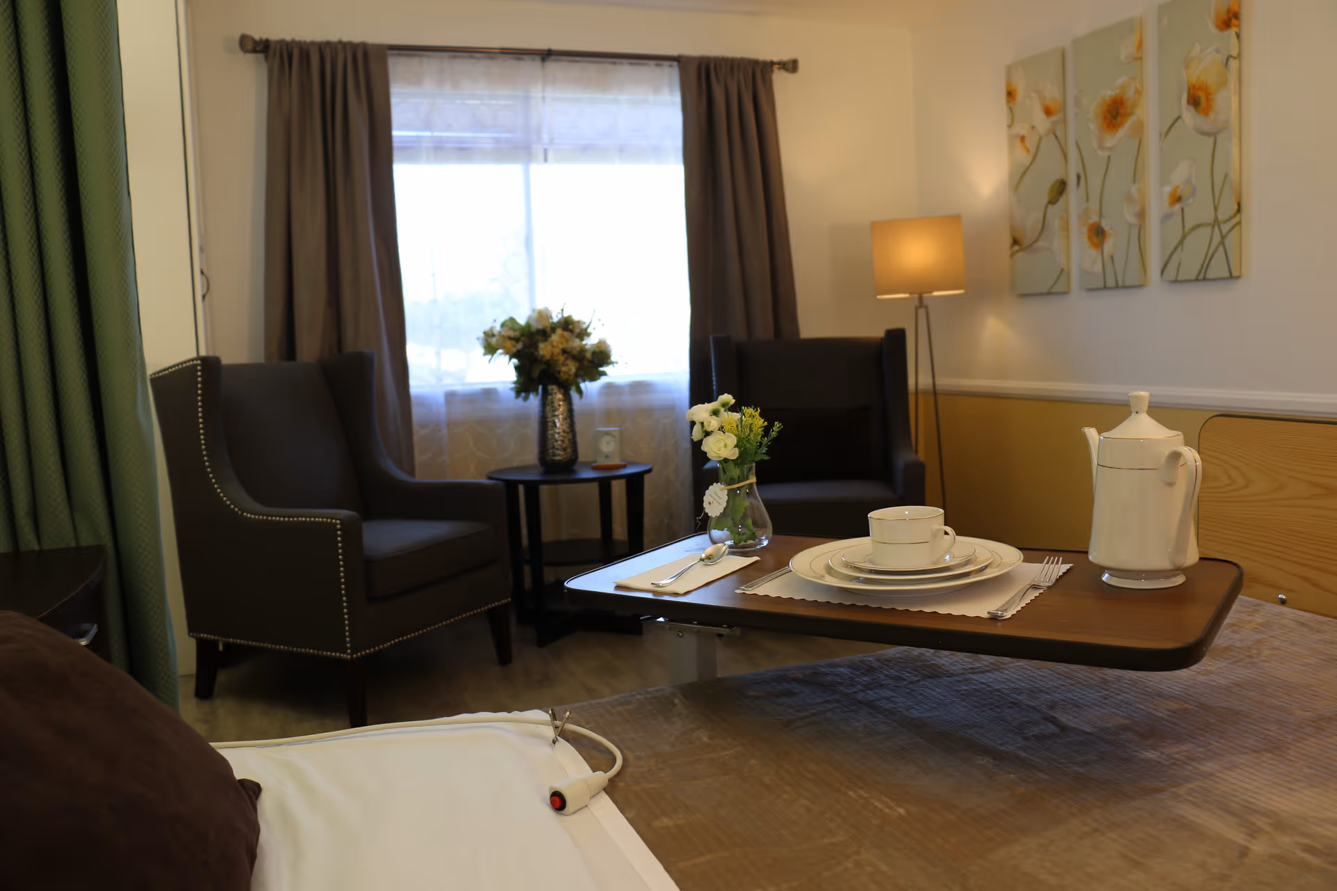 A cozy senior care bedroom with two armchairs by a window and a meal tray set over the bed.