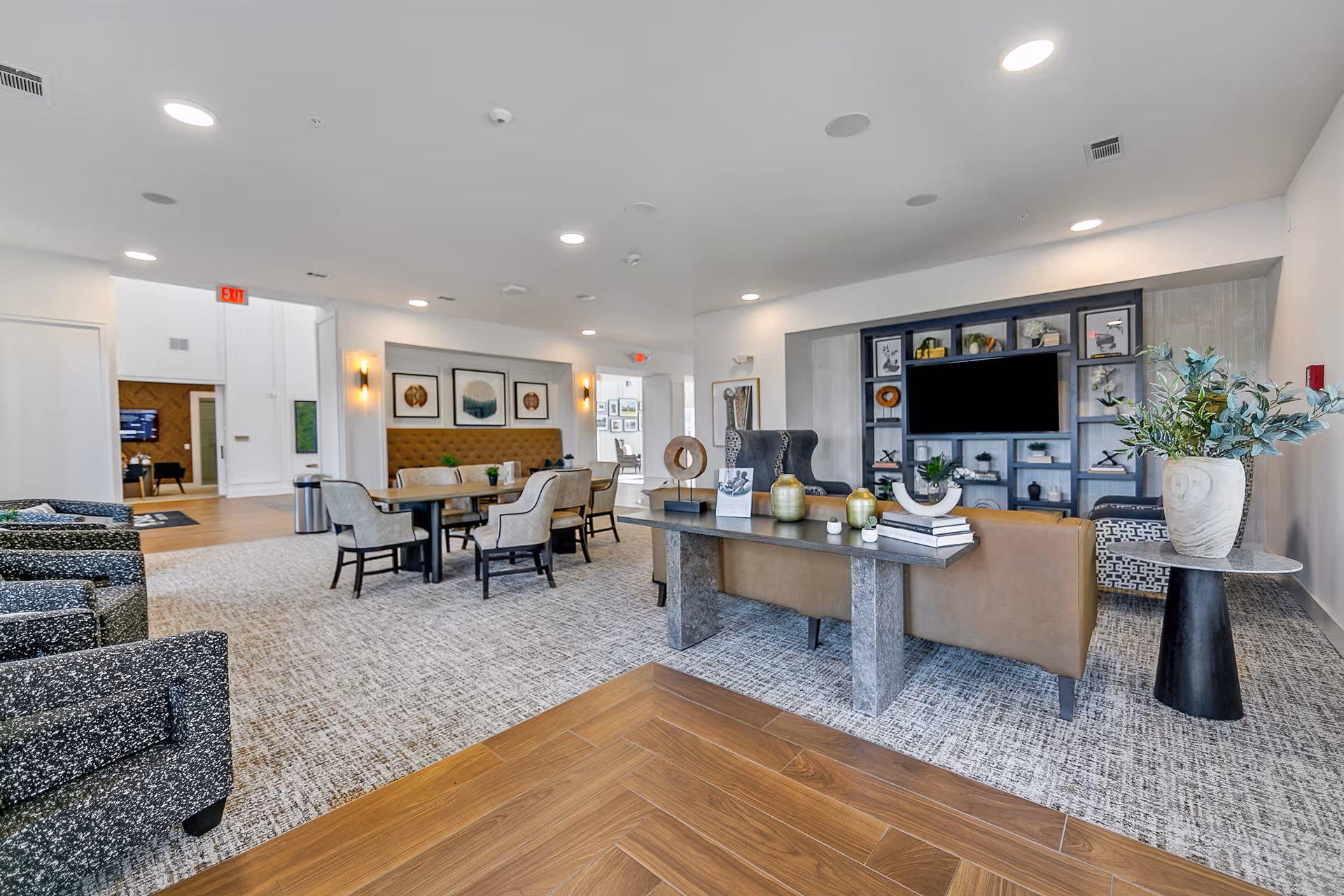 A spacious and modern living room area in a senior living facility featuring a large flat-screen TV mounted on a built-in shelving unit with decorative items. The room has a mix of seating including a brown leather sofa, patterned armchairs, and a dining table with upholstered chairs. The floor is a combination of wood and carpet, and the walls are decorated with framed artwork. The space is well-lit with recessed ceiling lights and wall sconces.