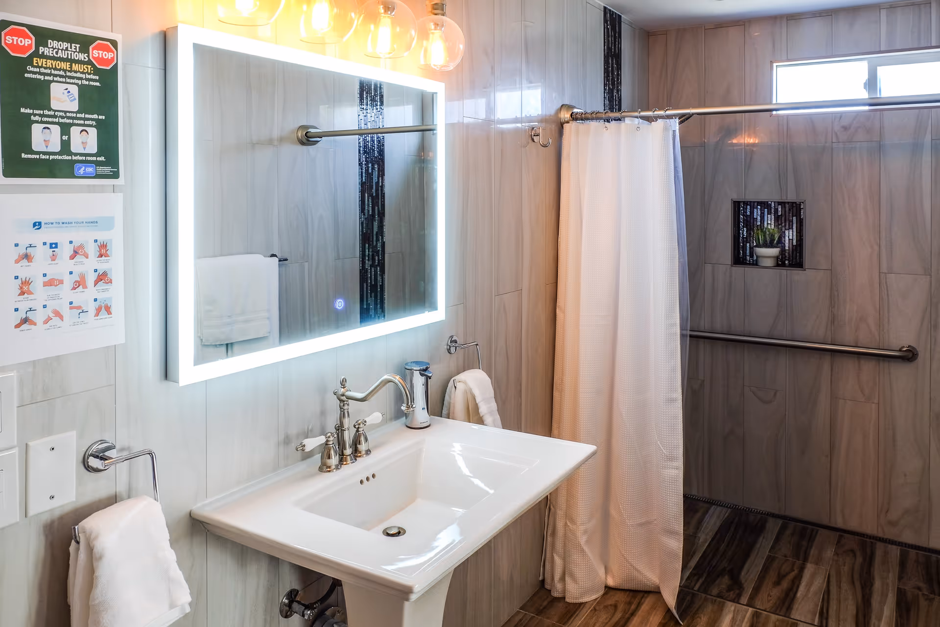A clean and modern bathroom with a white pedestal sink, a large illuminated mirror above it, and a shower area with a white curtain. The walls and floor have light-colored tiles with a vertical strip of dark mosaic tiles in the shower. There are handrails in the shower area and a small window letting in natural light. Two signs on the wall provide droplet precautions and hand washing instructions.