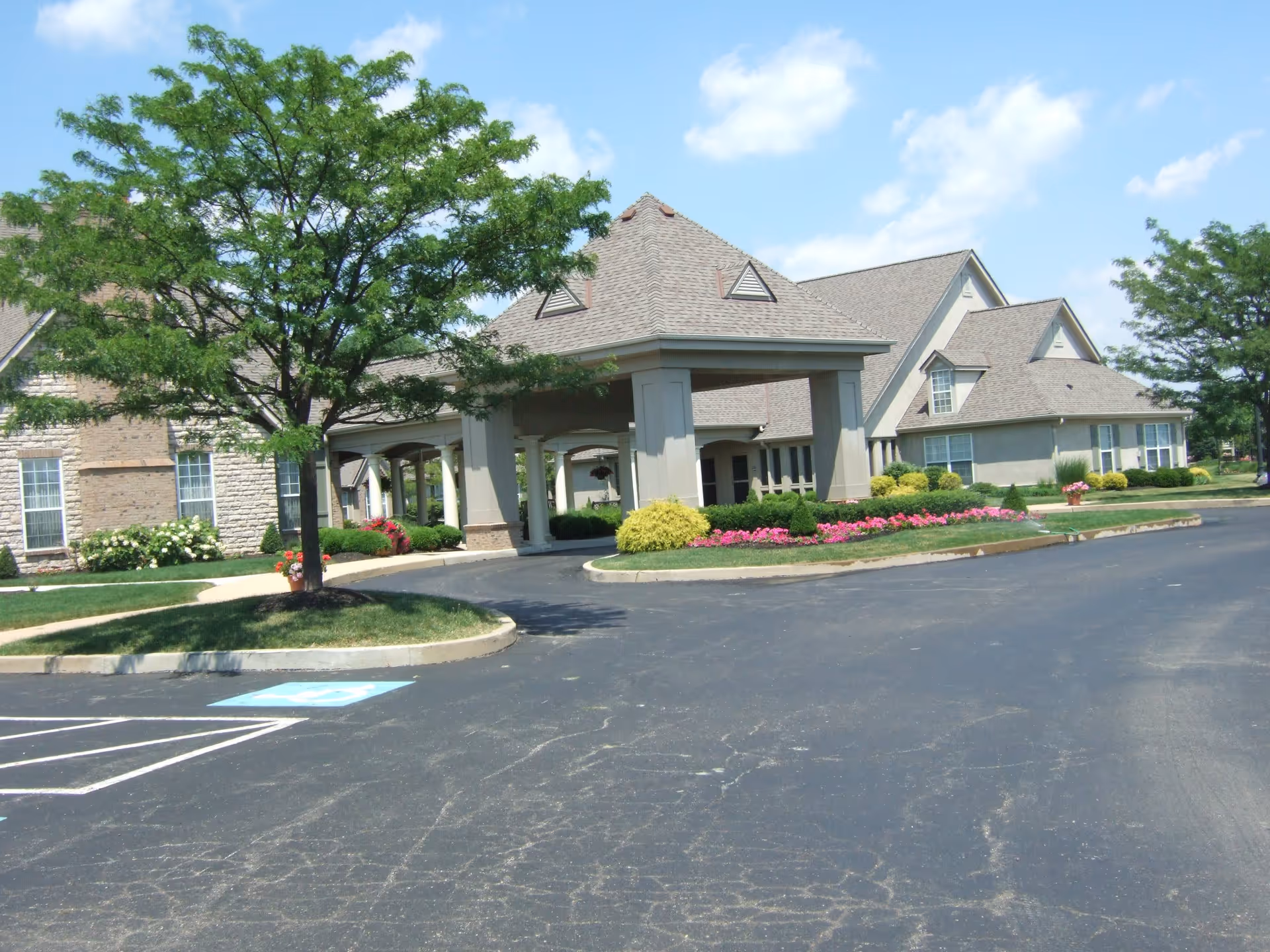 Front entrance of a senior living building with a covered drop-off canopy, landscaped flowerbeds, trees and a parking lot with a handicapped space.