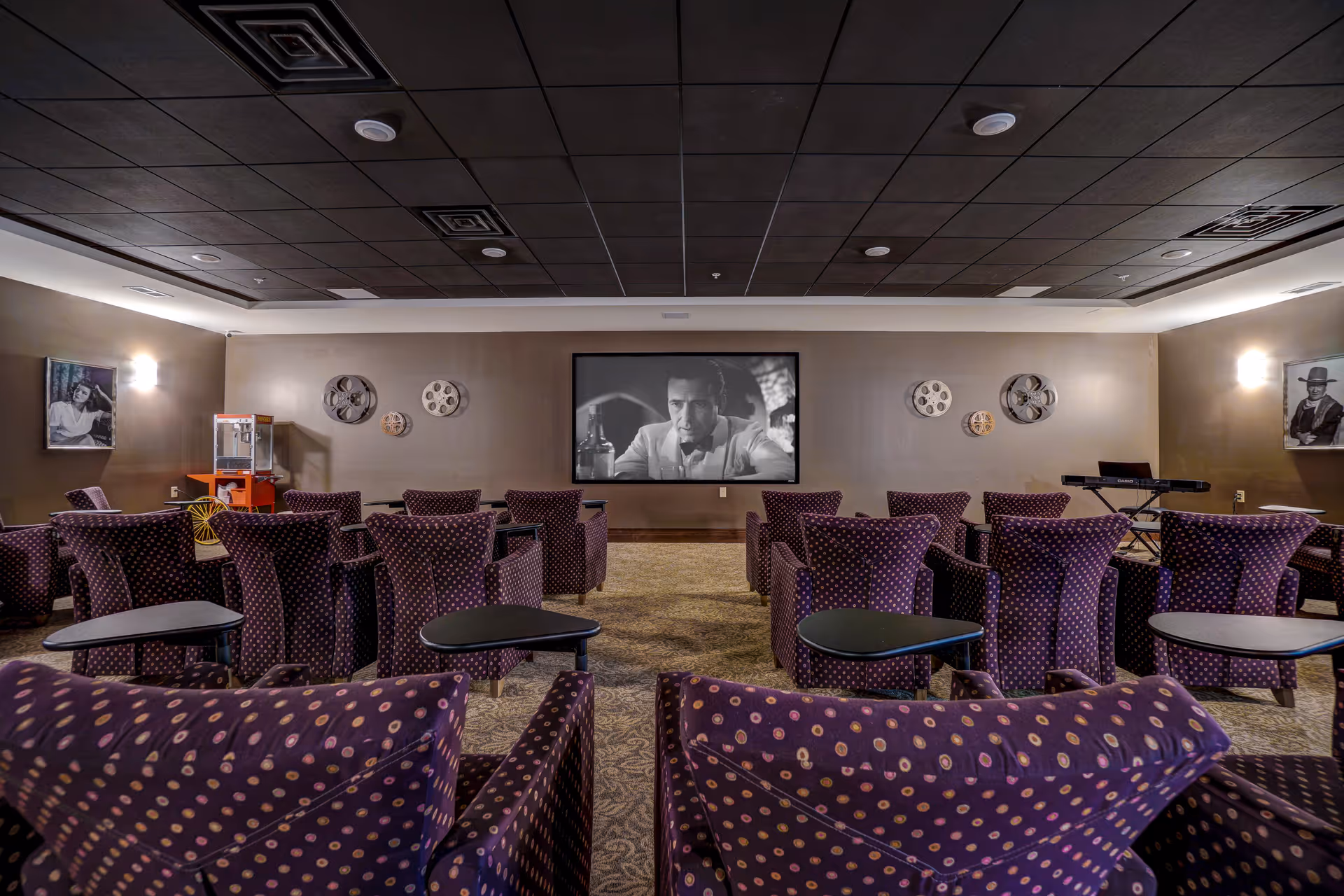 A cozy movie theater room with multiple rows of purple armchairs featuring a polka dot pattern. A large screen at the front displays a black and white film. The walls are decorated with vintage film reels and black and white portraits. There is a popcorn machine on the left side and a keyboard on the right side of the room.