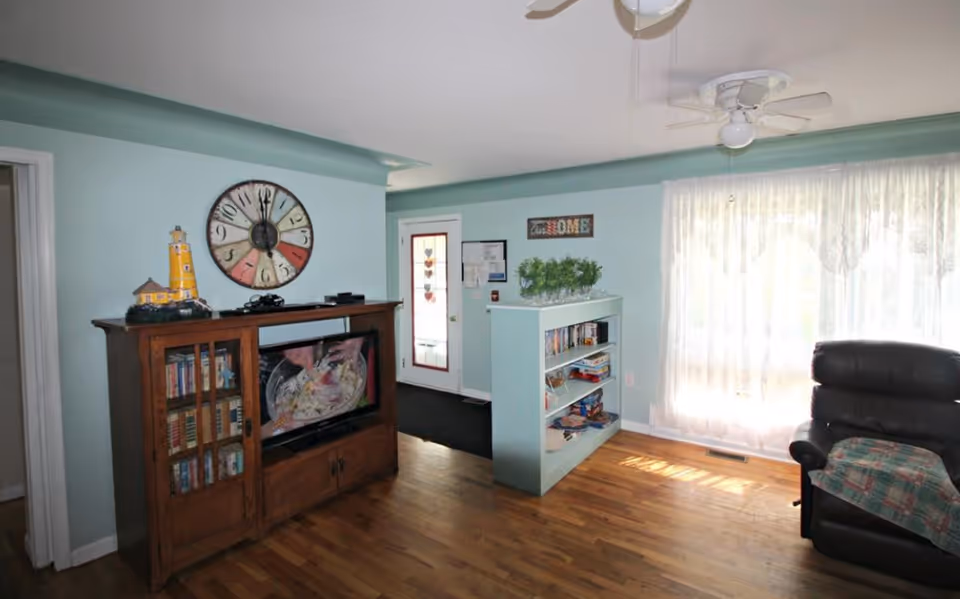 A cozy living room with light blue walls and hardwood floors. There is a wooden cabinet with glass doors holding books and a TV on top. Above the cabinet is a large colorful wall clock. To the right, a black recliner chair with a plaid blanket is positioned near a large window with sheer white curtains. A small bookshelf with plants and games is placed near a door with a glass panel decorated with heart shapes. A ceiling fan with lights hangs from the ceiling.