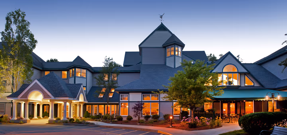 Front exterior of a large, well‑lit senior living facility at dusk with columns, landscaped grounds, and many illuminated windows.