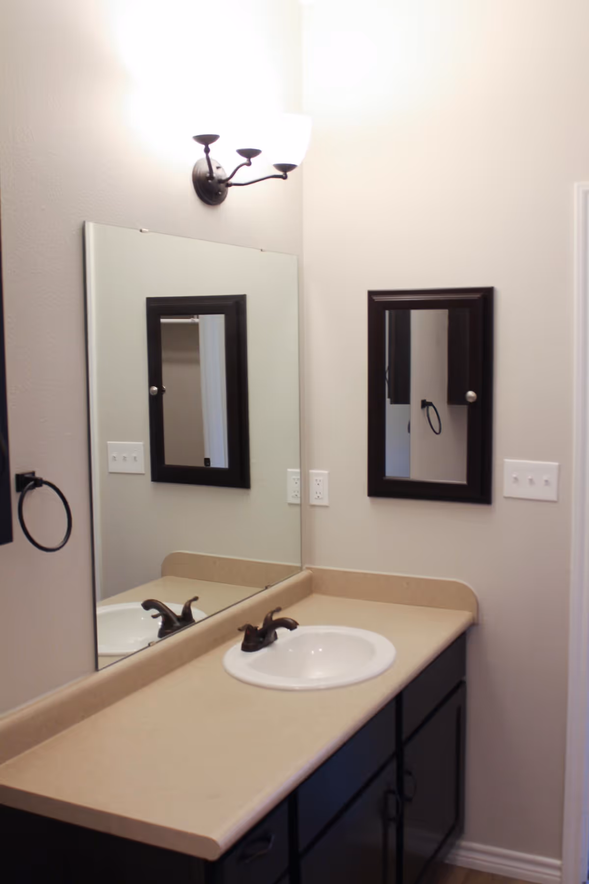 A bathroom vanity with a beige countertop, a white sink with a dark faucet, a large wall-mounted mirror, a smaller medicine cabinet with a dark frame, a wall-mounted light fixture above the mirror, and a towel ring on the left wall.