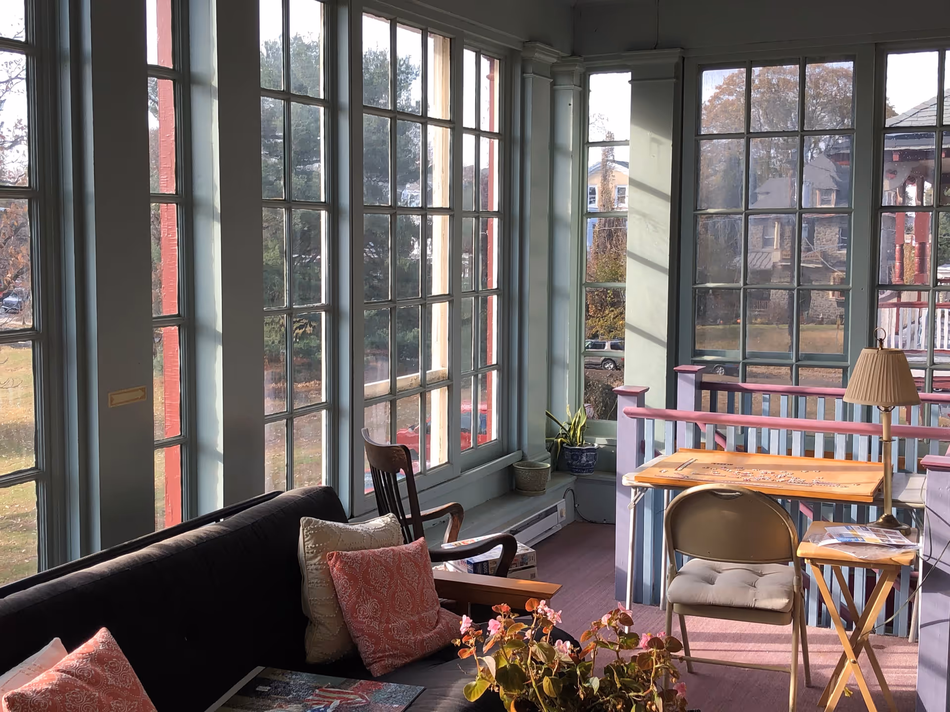 A sunlit enclosed porch area with large windows showing an outdoor view. The space includes a black cushioned bench with patterned pillows, a wooden chair, a small table with a lamp, and a folding chair. There are plants on the floor and a table with a puzzle on it.