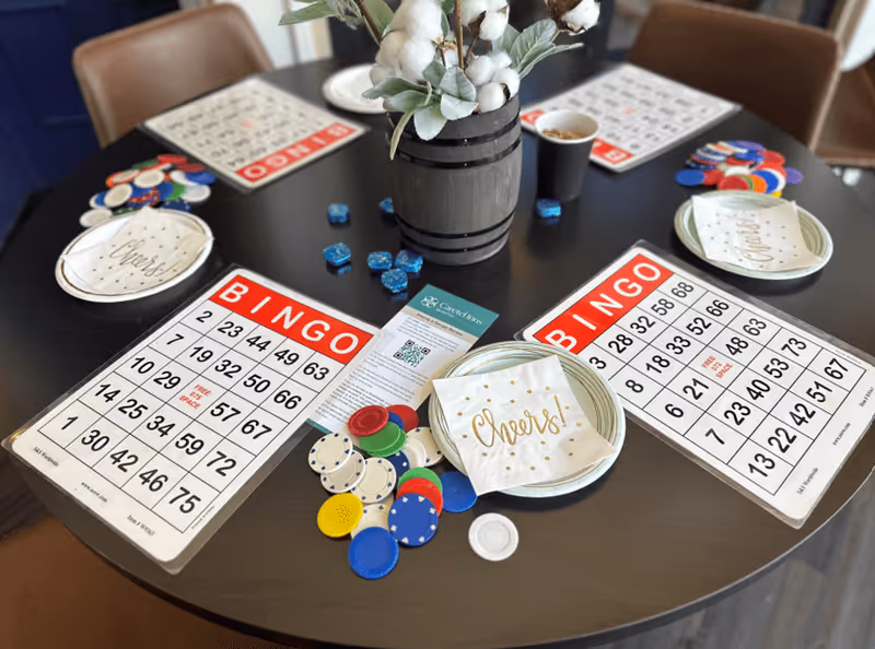 A round black table set up for a bingo game with bingo cards, colorful bingo chips, plates with napkins that say 'Cheers!', a cup, and a small vase with cotton plant decoration in the center.