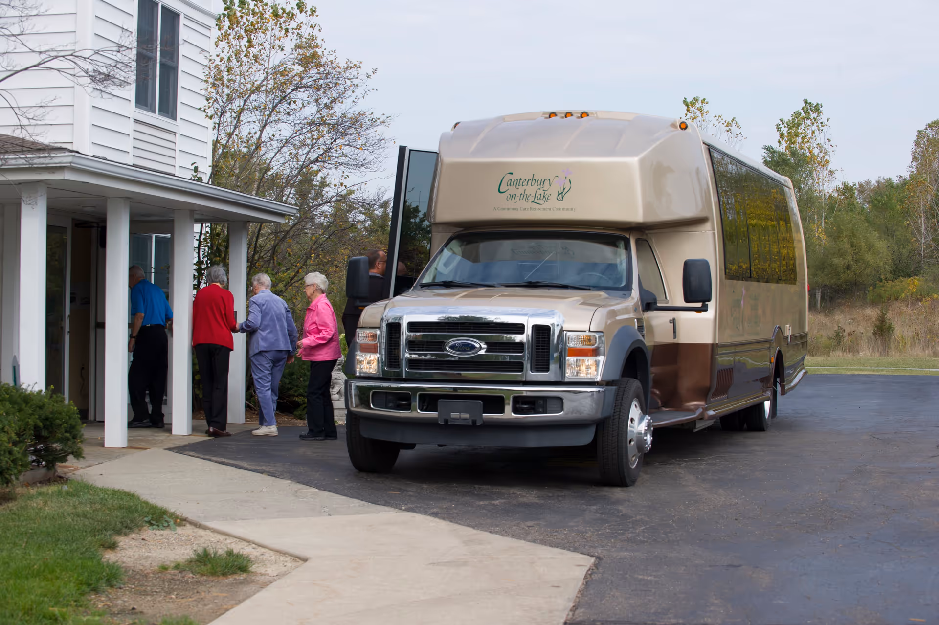 A group of elderly people entering a building next to a beige and brown shuttle bus with the logo 'Canterbury on the Lake' parked on a driveway. The scene is outdoors with some greenery and trees in the background.