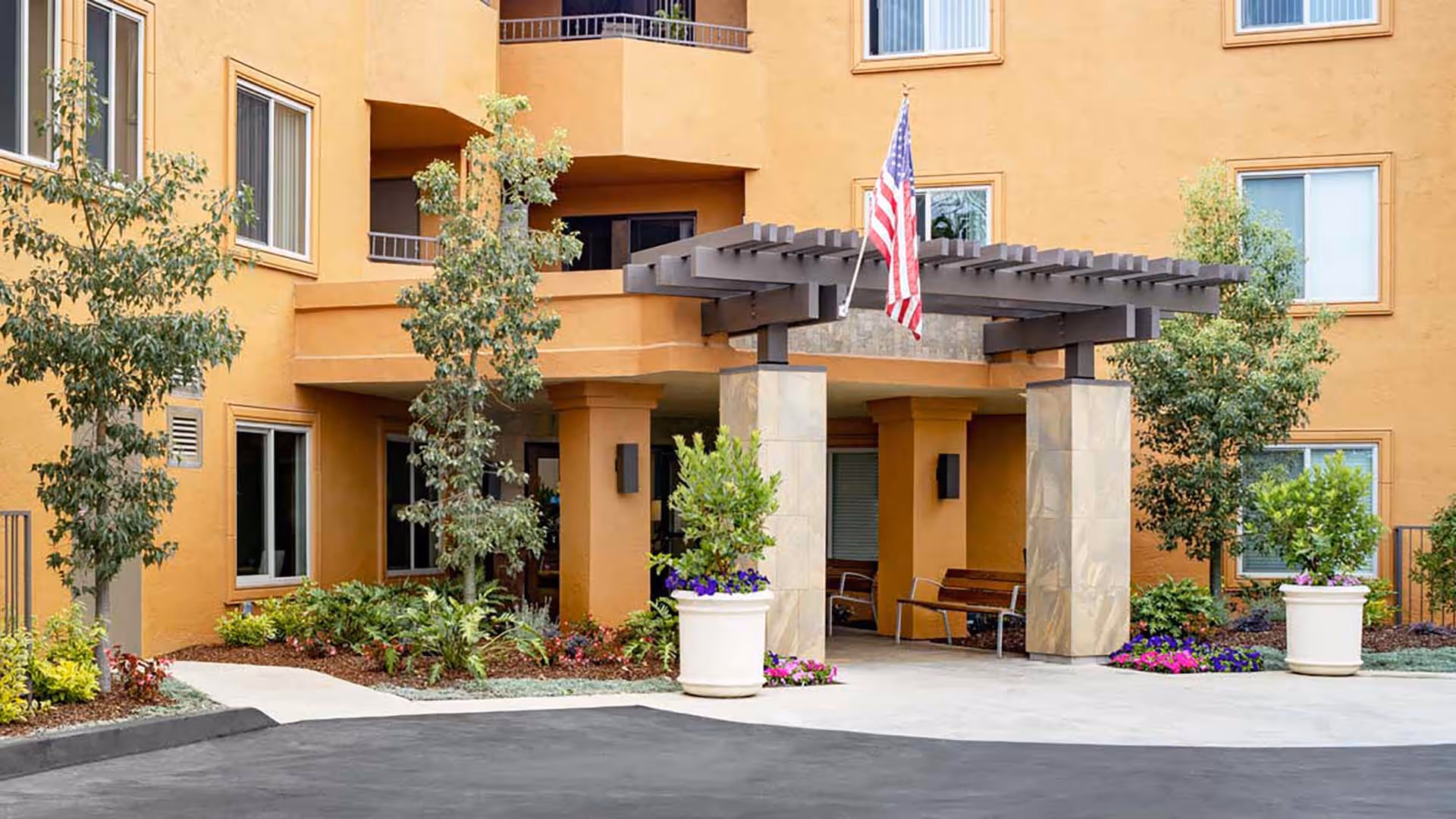 Entrance of a senior living facility with a tan exterior, a pergola supported by stone pillars, an American flag hanging from the pergola, landscaped greenery including trees and flowers, and benches near the entrance.