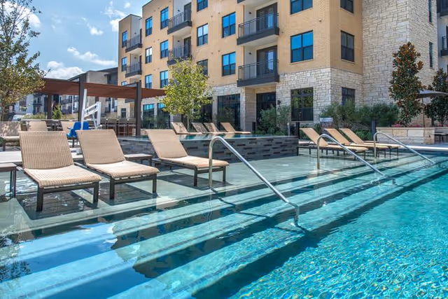 Outdoor swimming pool area with several lounge chairs partially submerged in shallow water, surrounded by a multi-story residential building with balconies, trees, and clear blue sky.