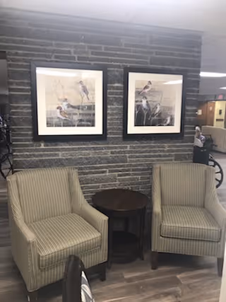 Two upholstered armchairs flank a round side table against a textured interior wall with two framed bird prints.