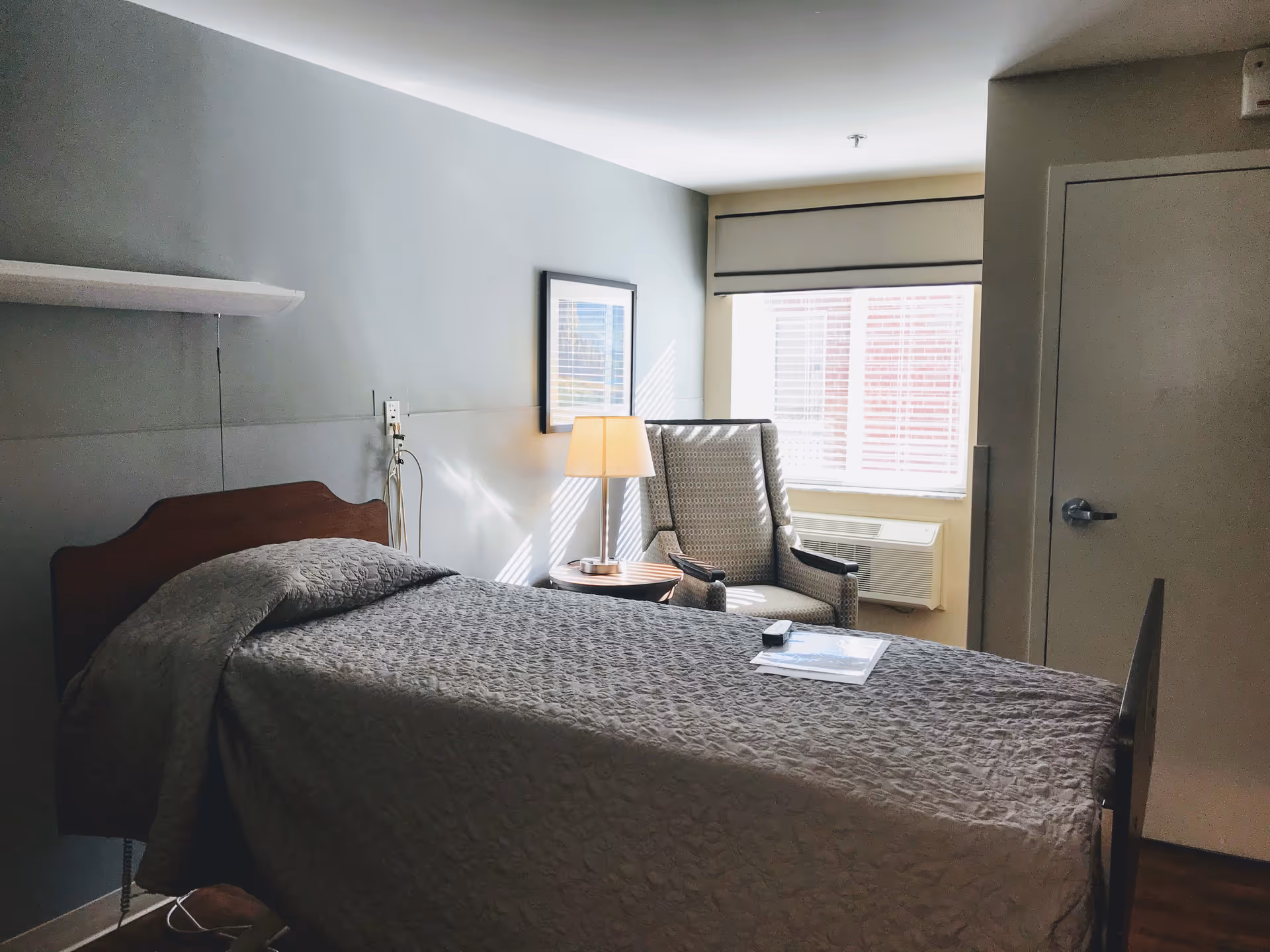 A simple nursing facility bedroom with a single bed covered in a gray quilt, a wooden headboard, a small round side table with a lamp, a patterned armchair near a window with blinds, and a closed door on the right side.