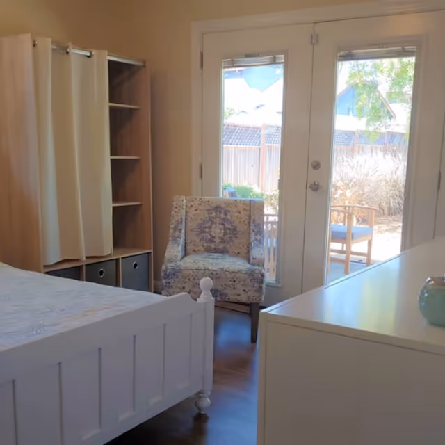 A bright bedroom featuring a white wooden bed frame with a light-colored bedspread, a patterned armchair, a wooden shelving unit with a curtain, and a white dresser. French doors with glass panes open to an outdoor patio area with chairs and plants visible outside.