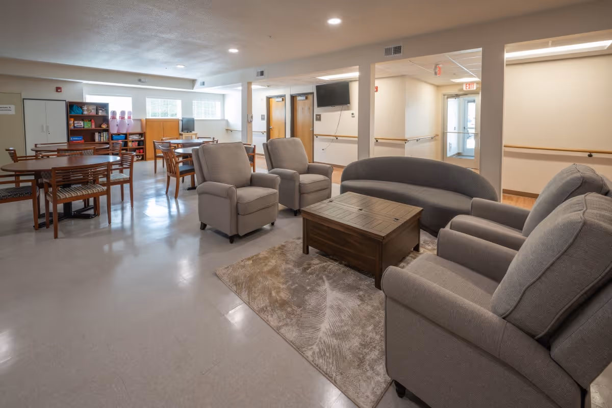 Communal senior living lounge with armchairs, a sofa, a coffee table and several dining tables in a bright open room.