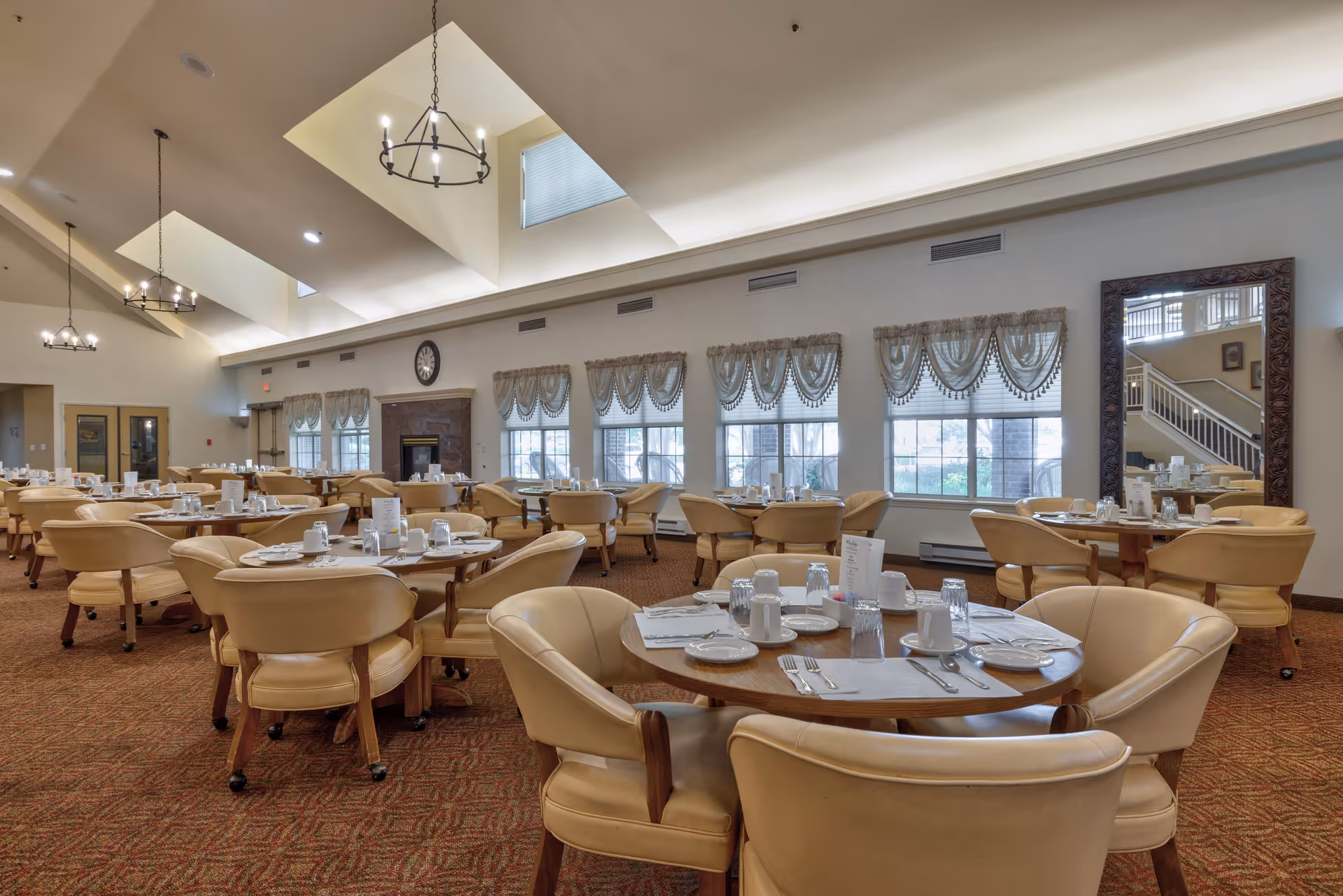 Spacious dining room with round tables set for meals, beige cushioned chairs, large windows with decorative curtains, high vaulted ceiling with chandeliers, and a large mirror on the right wall.