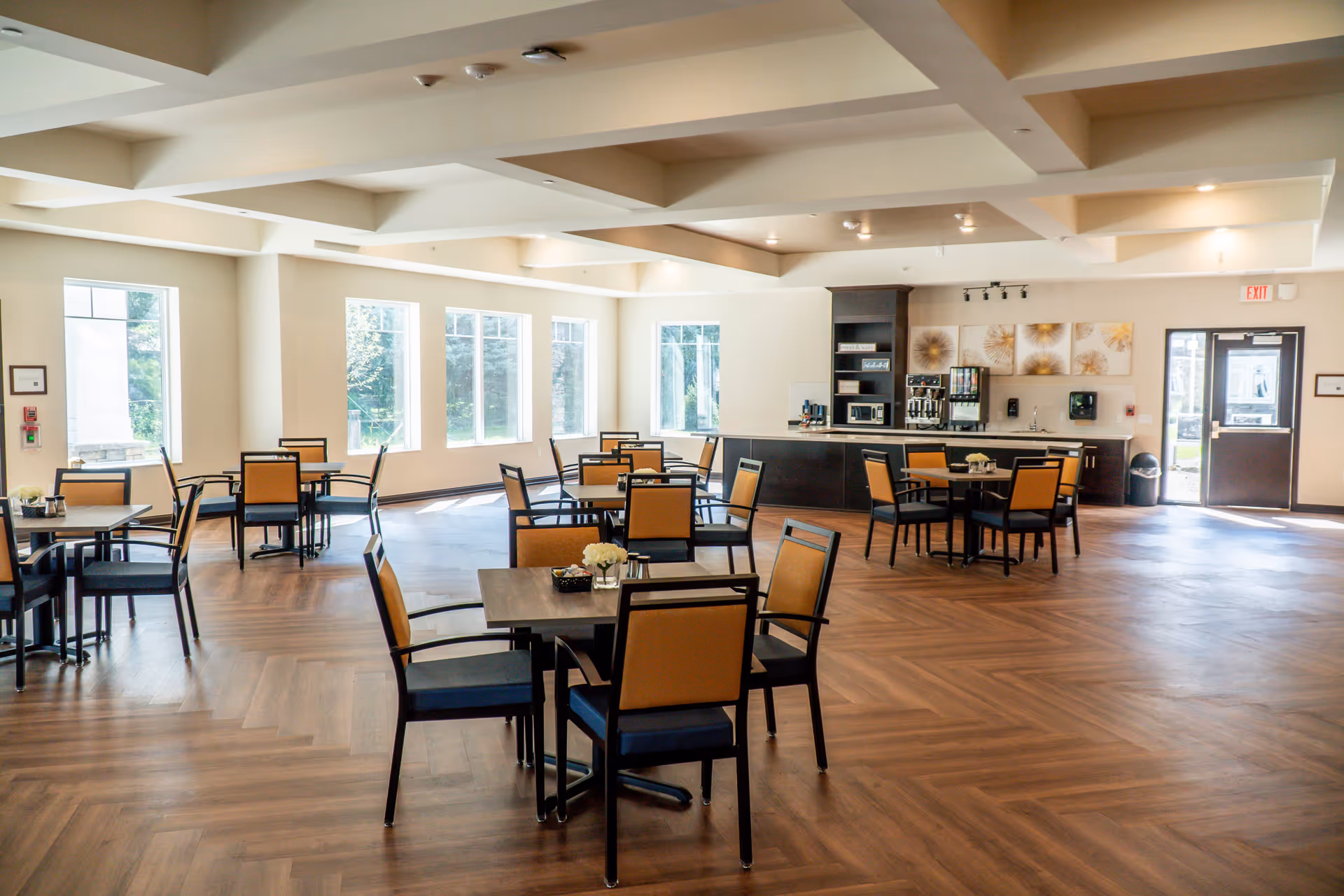 A bright and spacious dining room with multiple tables and chairs arranged neatly. The room features large windows allowing natural light to fill the space, a coffee and beverage station against the far wall, and modern decor with light-colored walls and wooden flooring.