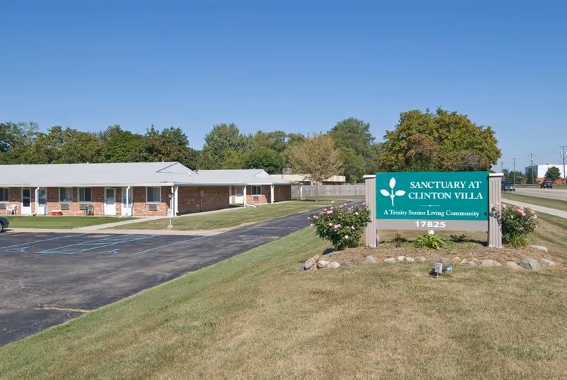 Exterior view of Sanctuary at Clinton Villa senior living community showing a single-story brick building with white doors and windows, a parking lot with marked spaces, and a green sign with white text reading 'Sanctuary at Clinton Villa, A Trinity Senior Living Community, 17825' surrounded by grass and small bushes under a clear blue sky.