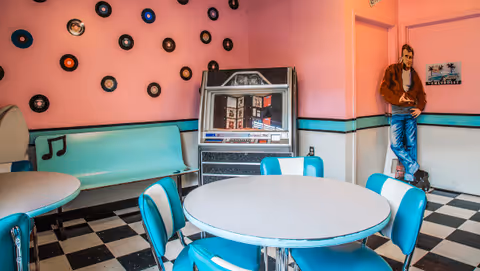A retro-style room with pink walls decorated with vinyl records and musical notes. There is a jukebox against the wall, a turquoise bench, and a round table surrounded by turquoise and white chairs. A life-sized cutout of a man in casual clothes leans against the wall near a door.